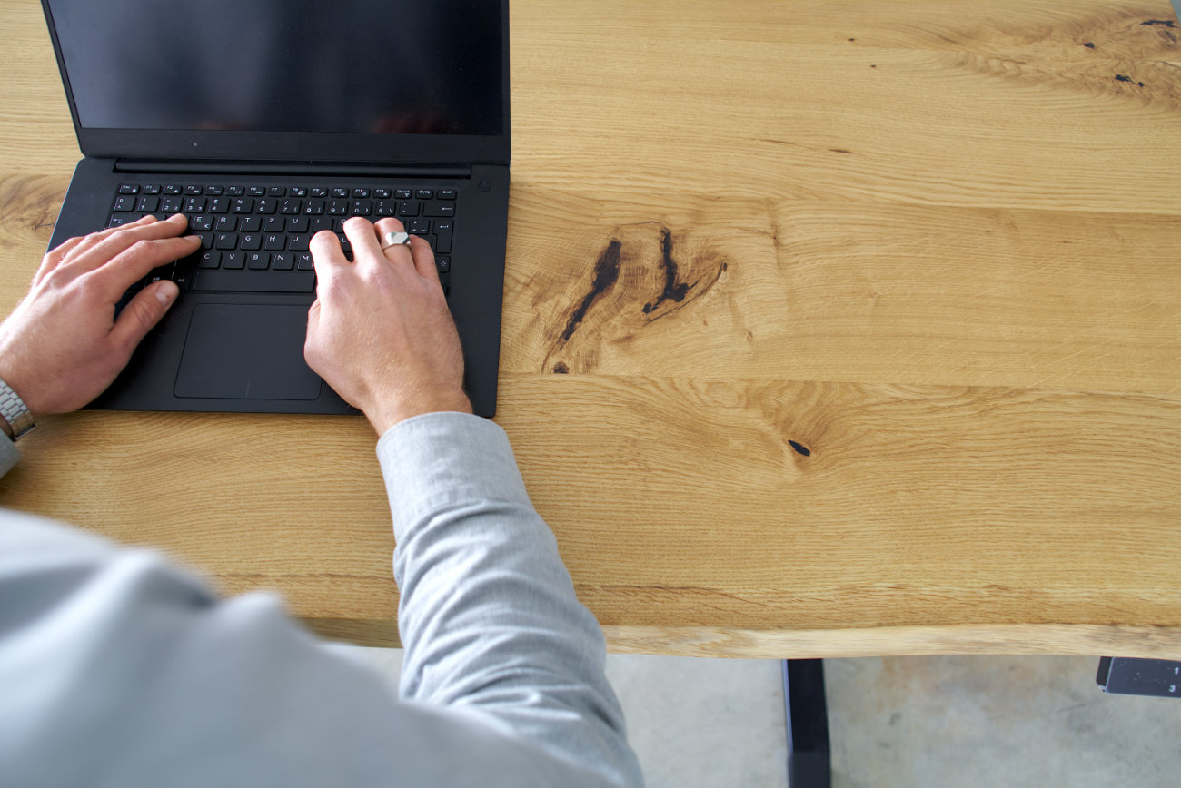 junger mann hinter einem höhenverstellbarem schreibtisch aus massivholz eiche mit baumkante darauf laptop und kaffeetasse